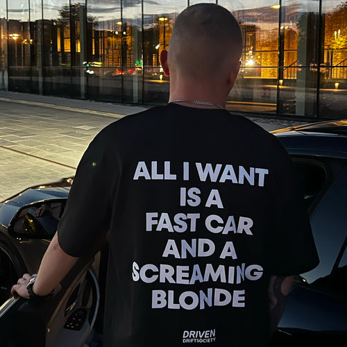 A man viewed from behind wearing a black t-shirt with bold white text that reads 'ALL I WANT IS A FAST CAR AND A SCREAMING BLONDE'. The shirt also features the 'DRIVEN DRIFT SOCIETY' logo at the bottom.