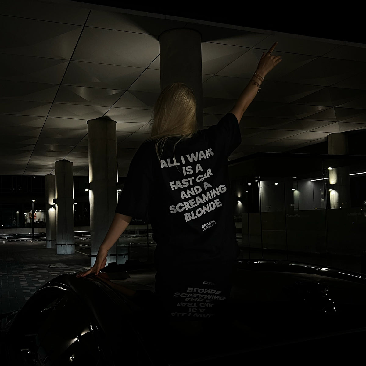 A girl sits on a car in parking space at night, viewed from behind, wearing a black graphic T-shirt with bold white text on the back. The shirt reads, 'ALL I WANT IS A FAST CAR AND A SCREAMING BLONDE'.