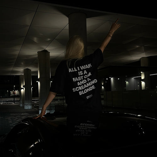 A girl sits on a car in parking space at night, viewed from behind, wearing a black graphic T-shirt with bold white text on the back. The shirt reads, 'ALL I WANT IS A FAST CAR AND A SCREAMING BLONDE'.