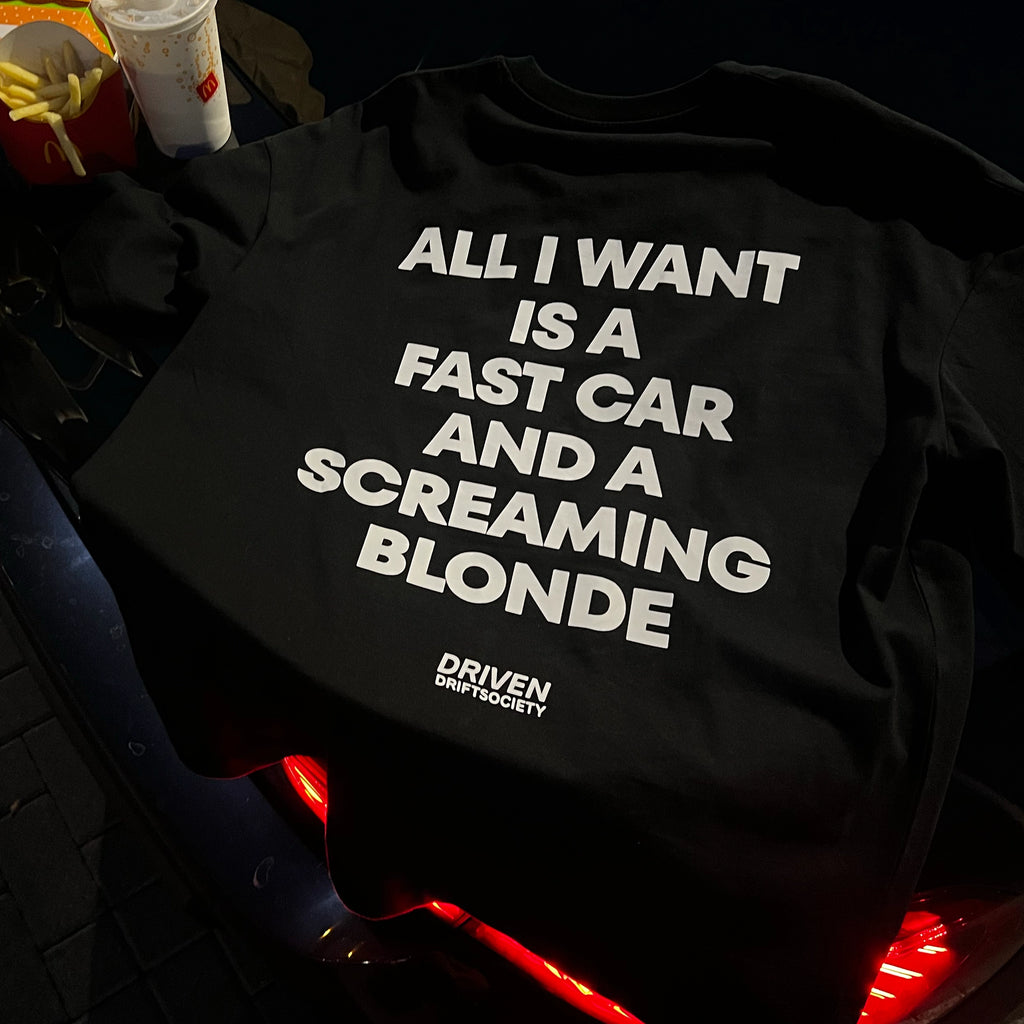 A black graphic t-shirt with bold white text reading 'ALL I WANT IS A FAST CAR AND A SCREAMING BLONDE' and a small 'DRIVEN DRIFT SOCIETY' logo, laid on a dark car surface with fast food props nearby.