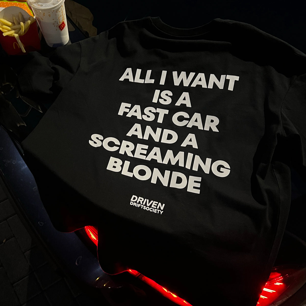 A black graphic t-shirt with bold white text reading 'ALL I WANT IS A FAST CAR AND A SCREAMING BLONDE' and a small 'DRIVEN DRIFT SOCIETY' logo, laid on a dark car surface with fast food props nearby.