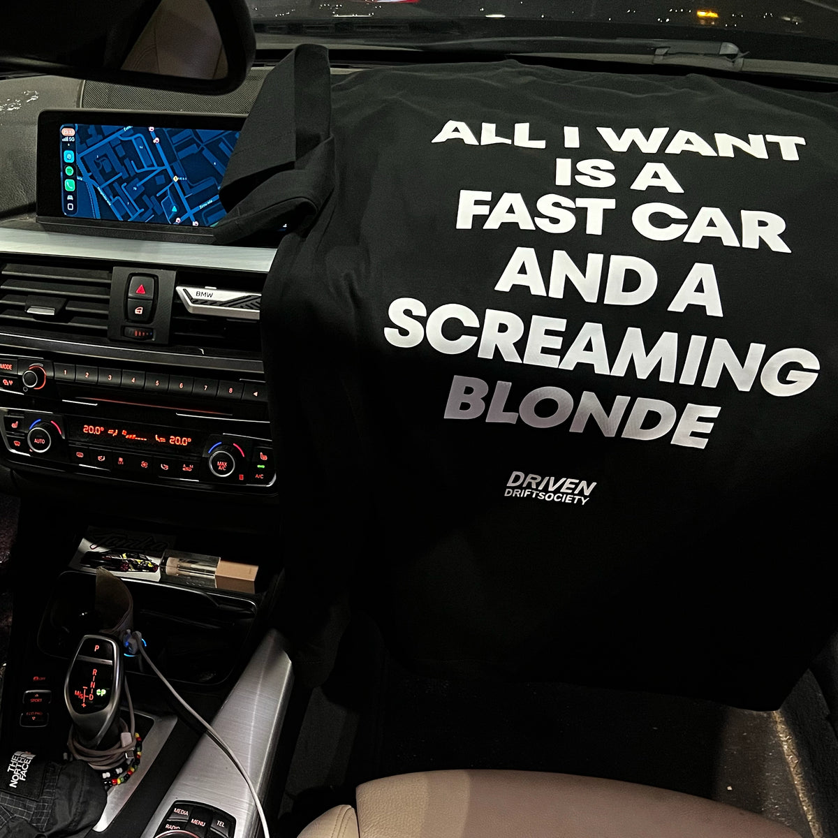 A black t-shirt with bold white text reading 'ALL I WANT IS A FAST CAR AND A SCREAMING BLONDE' and a small 'DRIVEN' logo shown placed on a car panel inside a vehicle interior.