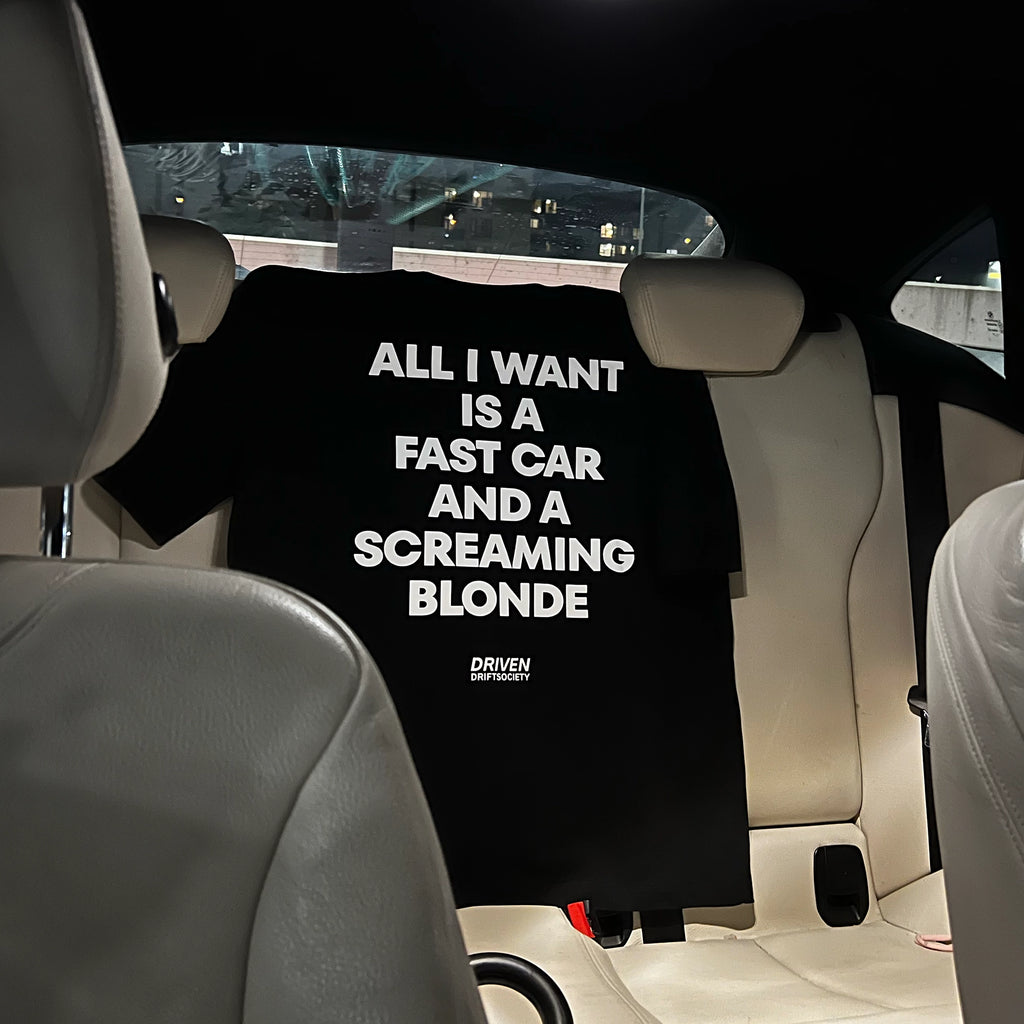 A black graphic t-shirt with bold white text reading 'ALL I WANT IS A FAST CAR AND A SCREAMING BLONDE' is draped over the back seat of a car. The shirt features a small 'DRIVEN DRIFT SOCIETY' logo at the bottom.