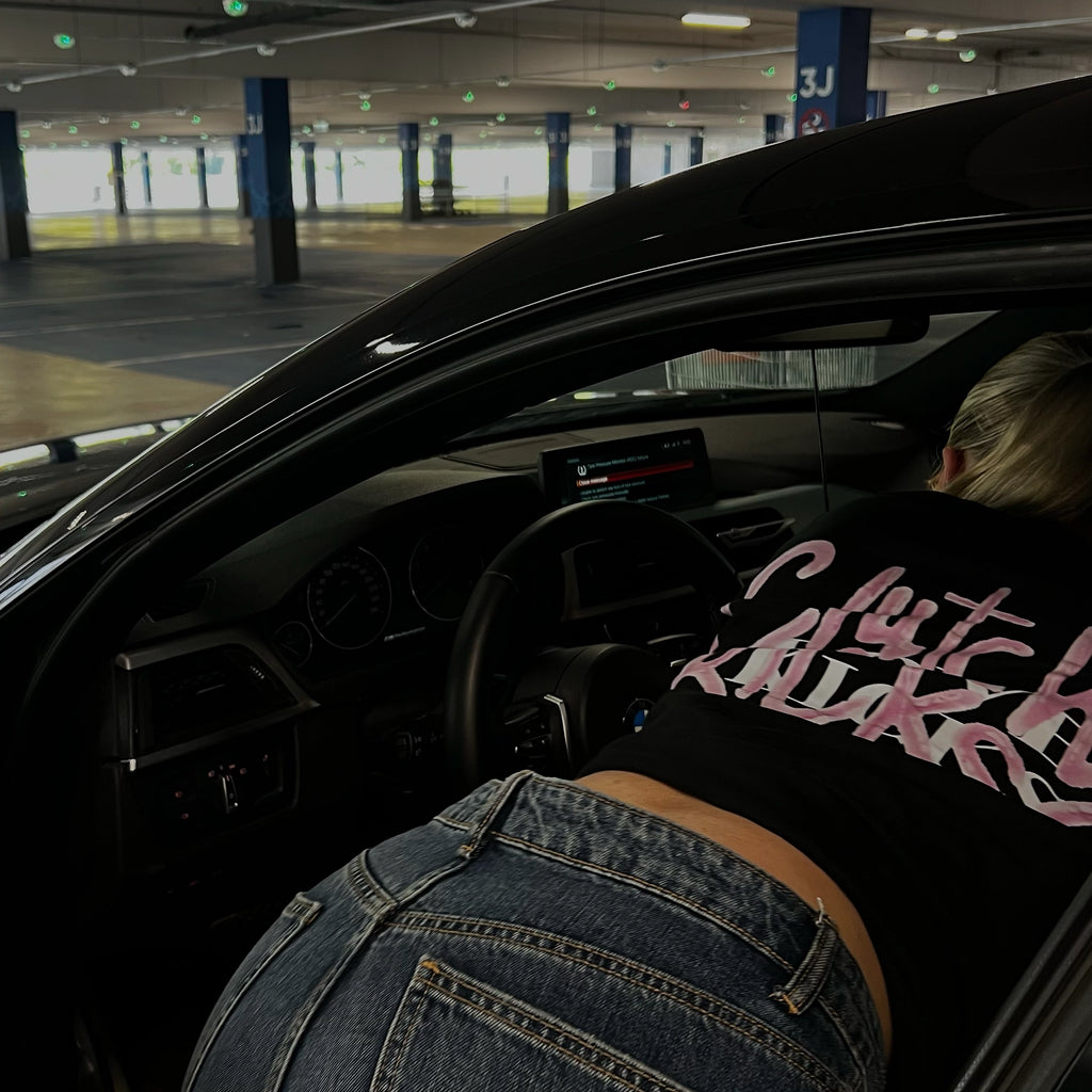 A person wearing a black t-shirt with pink cursive text and blue jeans leans into the open driver's side door of a black car in a parking garage.