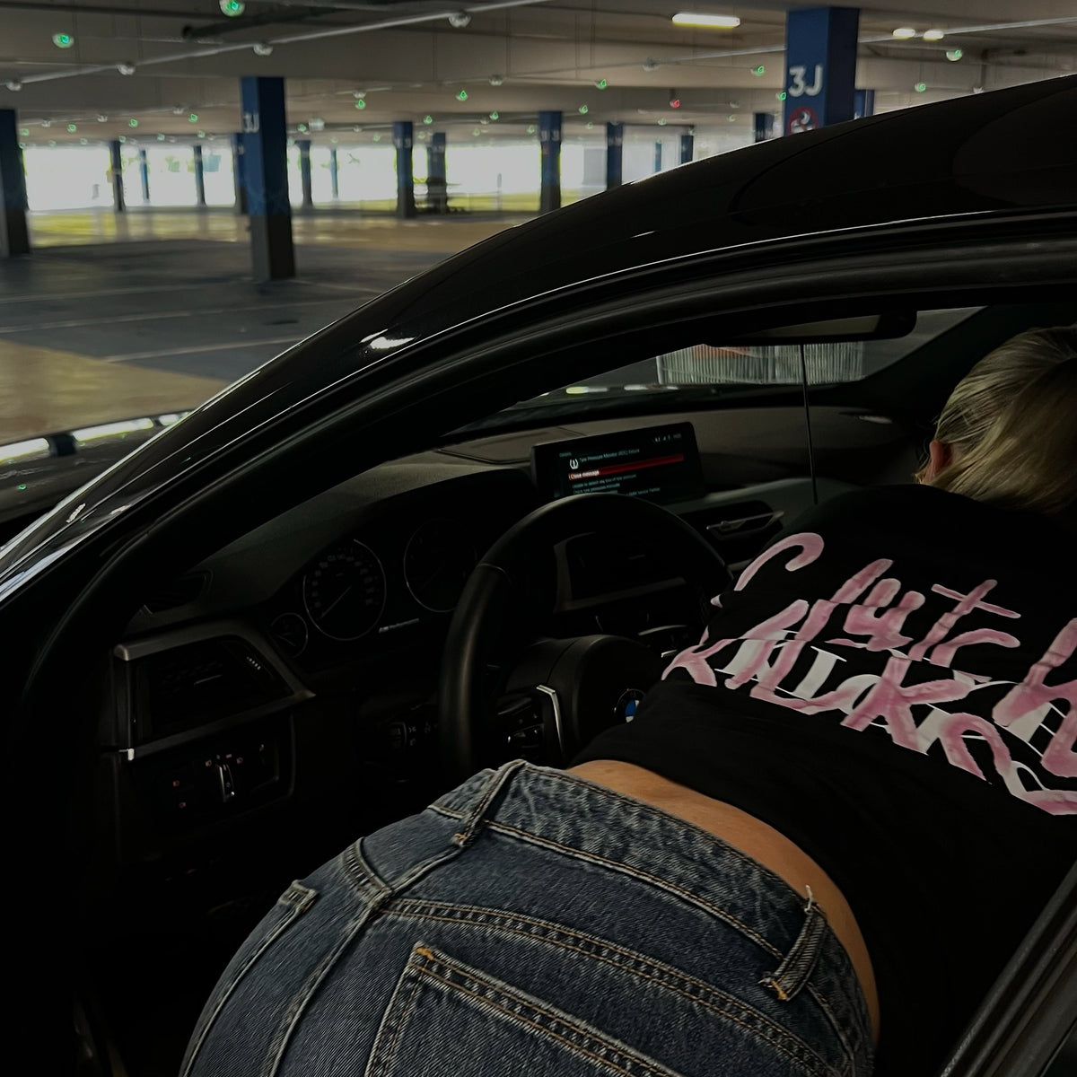 A person wearing a black t-shirt with pink cursive text and blue jeans leans into the open driver's side door of a black car in a parking garage.