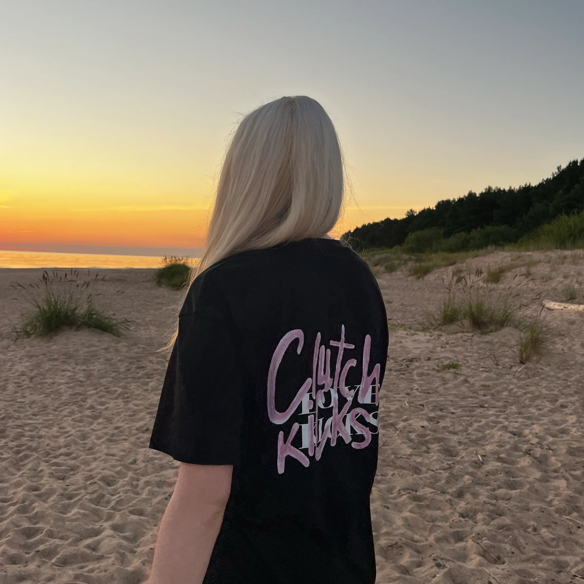 A person standing on a sandy beach at sunset, viewed from behind, wearing a black oversized T-shirt with a large pink and white graphic text on the back.