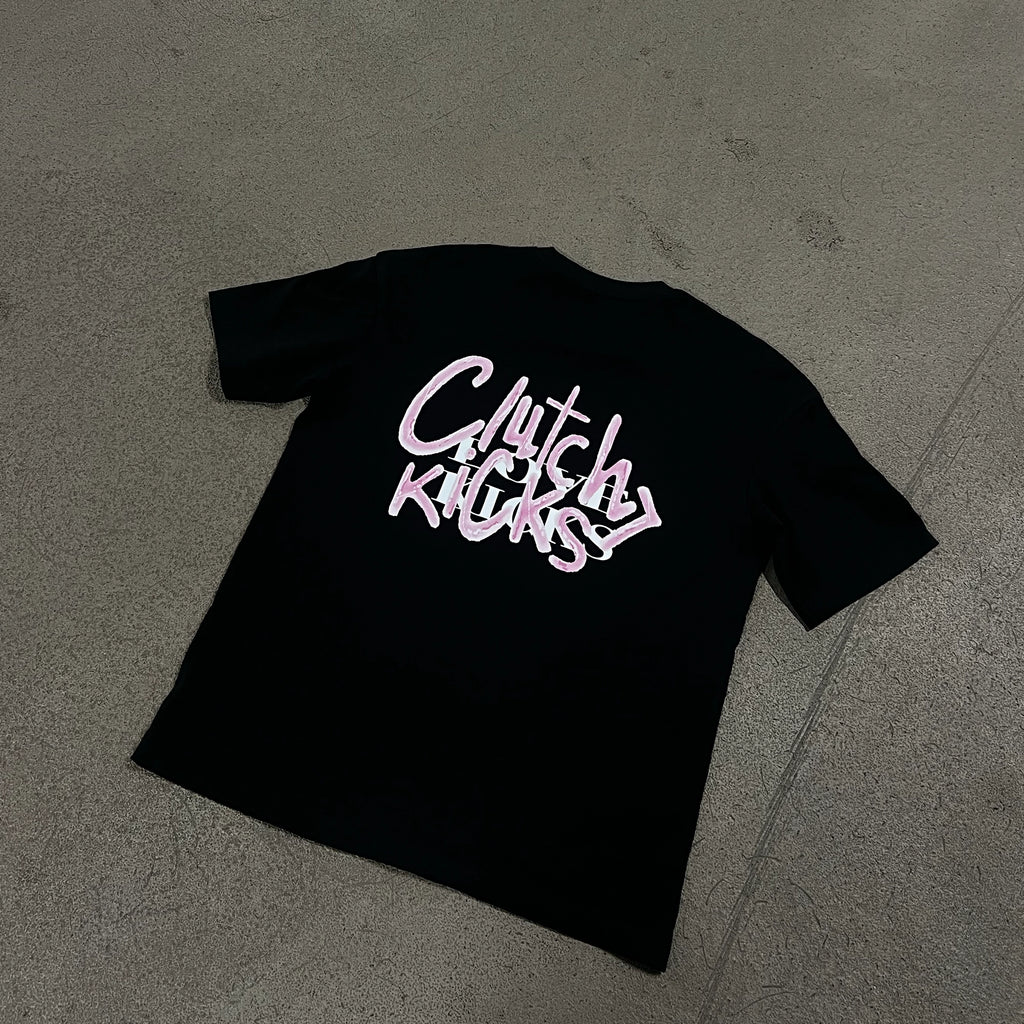 A black short-sleeve t-shirt laid flat on a gray concrete floor, featuring a stylized graphic on the back in white and pink that reads 'Clutch Kicks > Love Kicks'.
