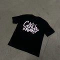 A black short-sleeve t-shirt laid flat on a gray concrete floor, featuring a stylized graphic on the back in white and pink that reads 'Clutch Kicks > Love Kicks'.
