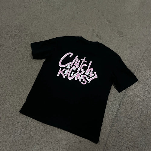 A black short-sleeve t-shirt laid flat on a gray concrete floor, featuring a stylized graphic on the back in white and pink that reads 'Clutch Kicks > Love Kicks'.