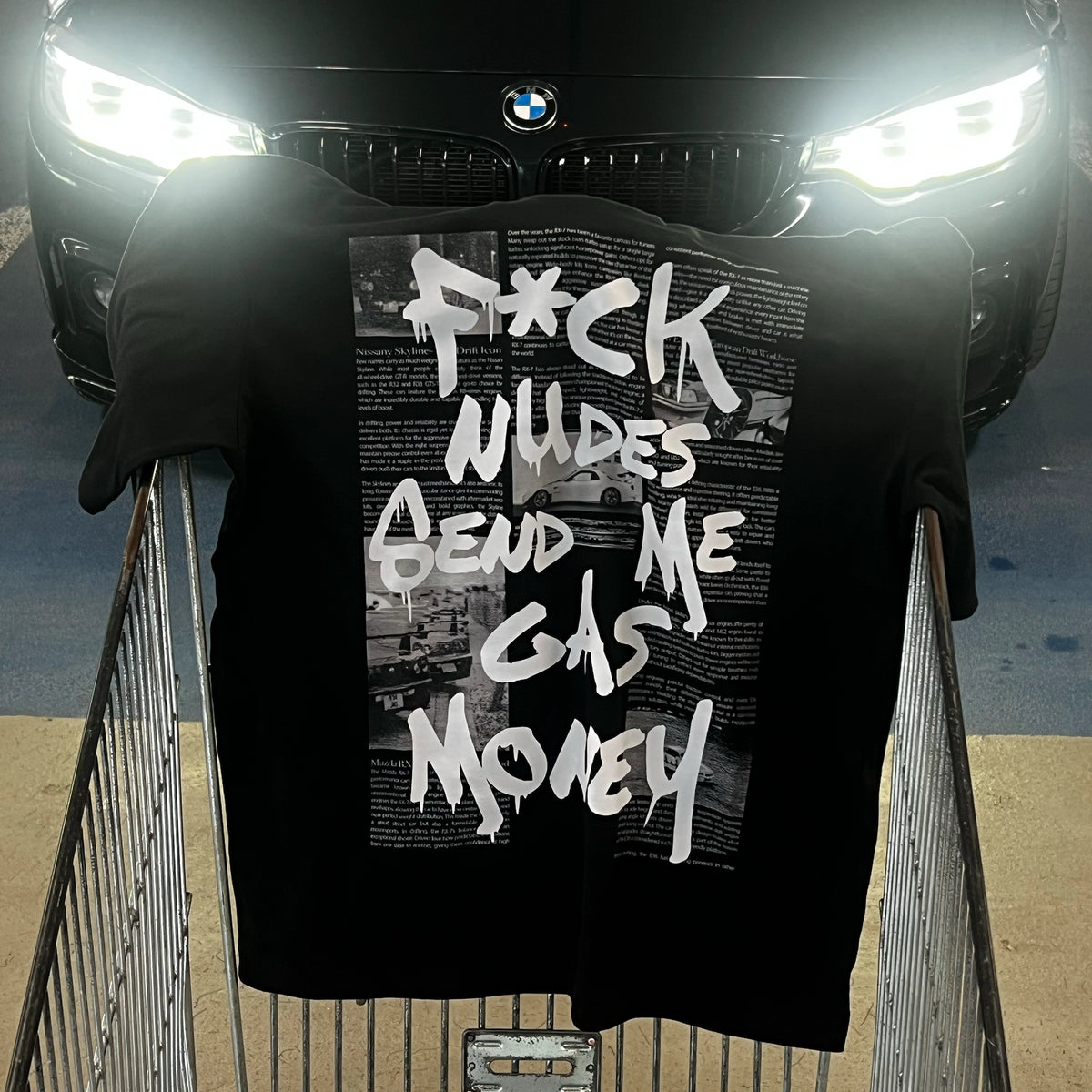 A black graphic t-shirt with bold white text reading 'F*CK NUDES SEND ME GAS MONEY' is displayed on a shopping cart in front of a car with bright headlights. The car grille and BMW logo are visible in the background.