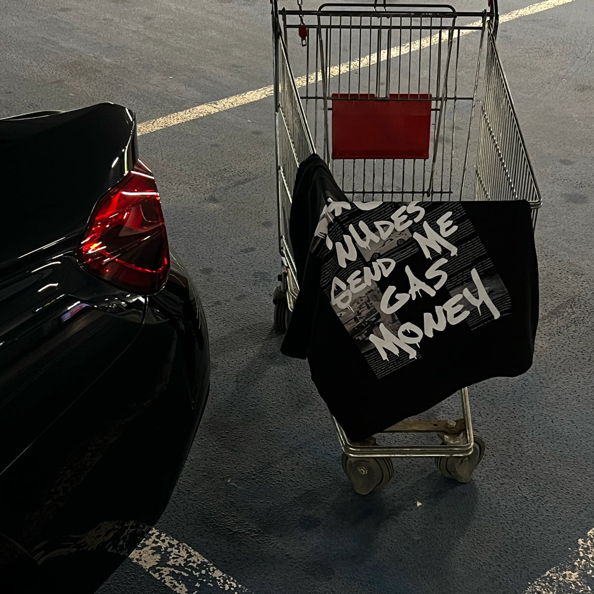 A black graphic T-shirt with white text reading 'F*CK NUDES SEND ME GAS MONEY' is draped over a metal shopping cart in a parking lot next to a black car.