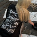 A person with long blonde hair seen from behind, wearing a black t-shirt with a large white graffiti-style graphic text print 'f*ck n*des send me gas money' on the back, standing on a cobblestone street.