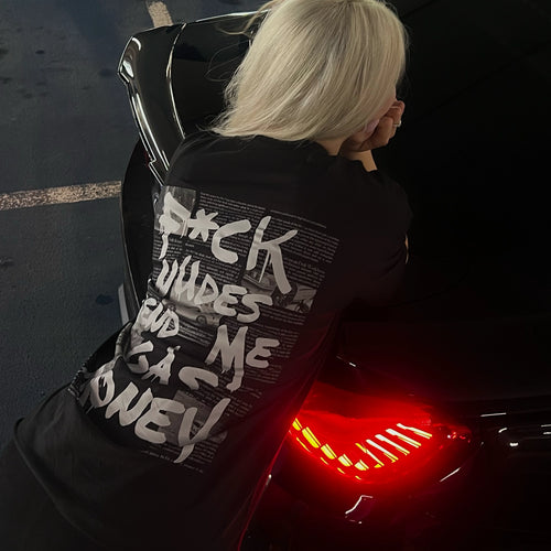 A person with platinum blonde hair seen from behind, leaning against a black car at night. The person is wearing a black t-shirt with a large white graphic text print  'f*ck n*des send me gas money' on the back.